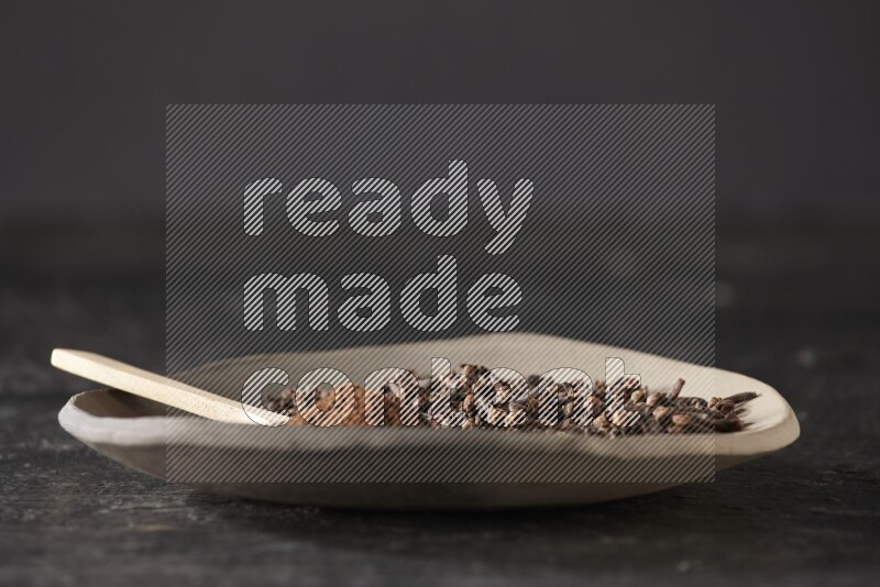 A Pottery plate full of cloves and a wooden spoon full of cloves powder on it on a textured black background