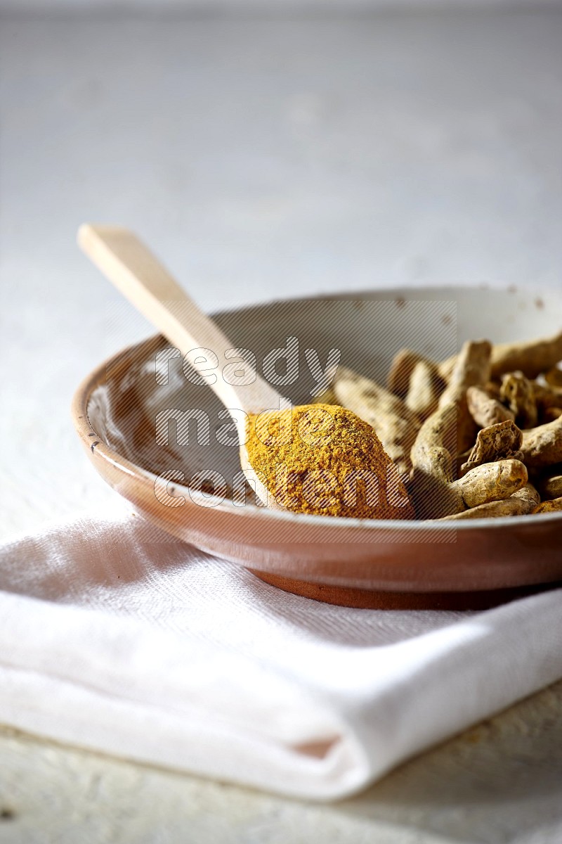 A plate filled with dried turmeric whole fingers and a wooden spoon full of turmeric powder on a textured white flooring