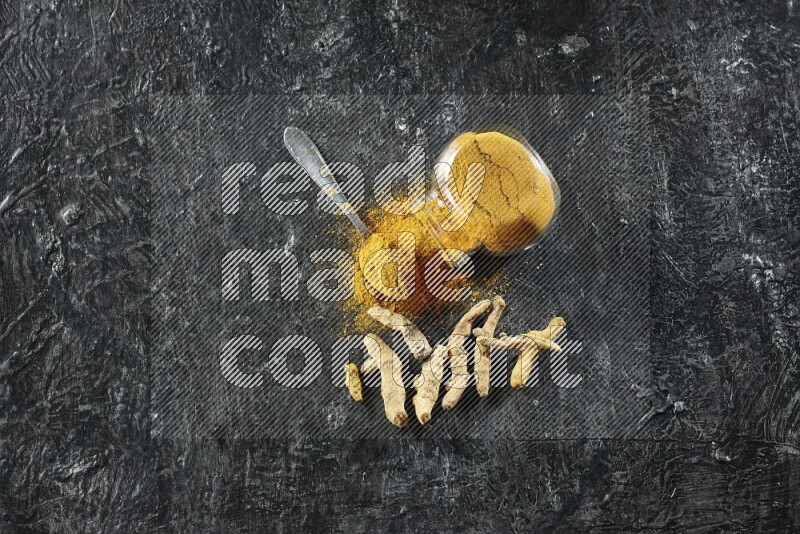 A flipped glass spice jar and metal spoon full of turmeric powder and powder spilled out of it with dried whole fingers on textured black flooring