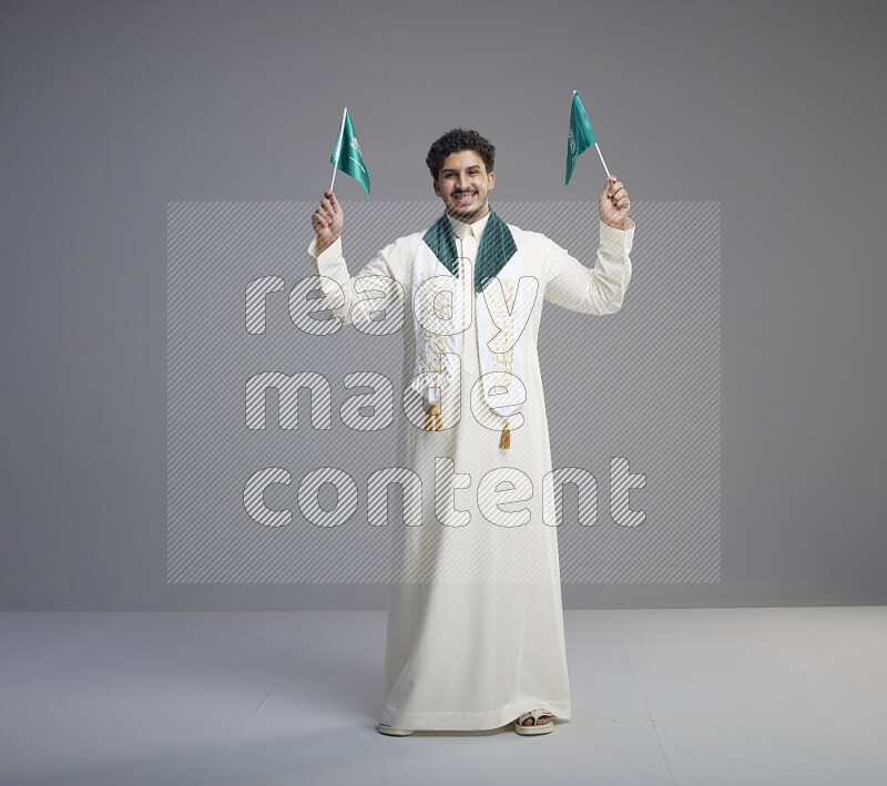 A Saudi man standing wearing thob and saudi flag scarf and holding small saudi flag on gray background