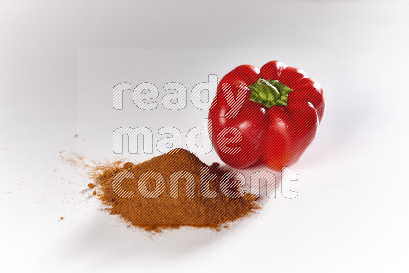 A bunch of ground paprika powder with a red bell pepper beside it on white background