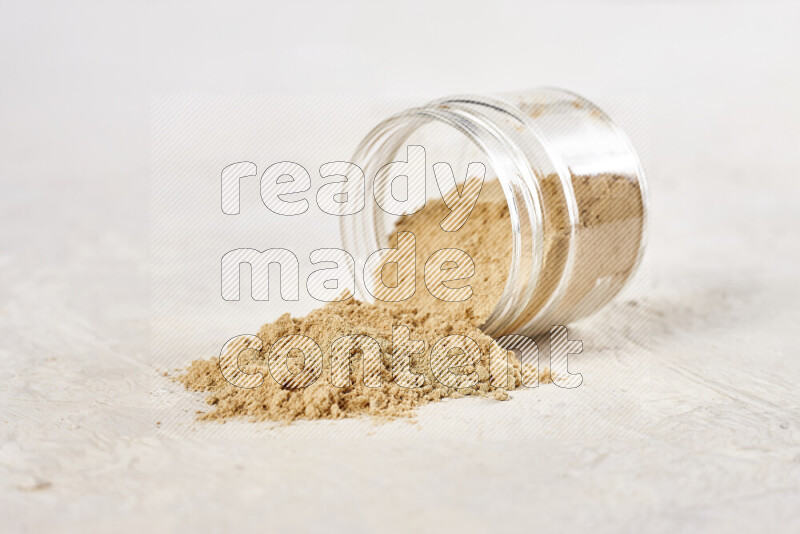 A glass jar full of ground ginger powder flipped with some spilling powder on white background