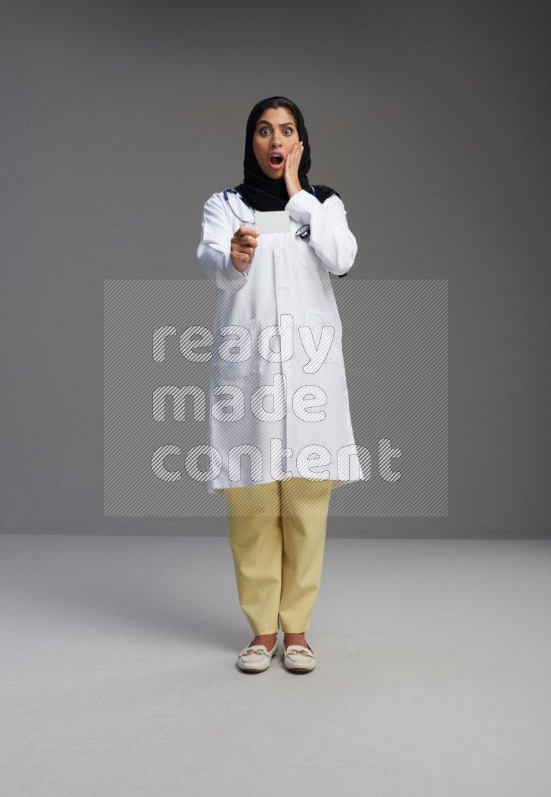 Saudi woman wearing lab coat with stethoscope standing holding ATM card on Gray background