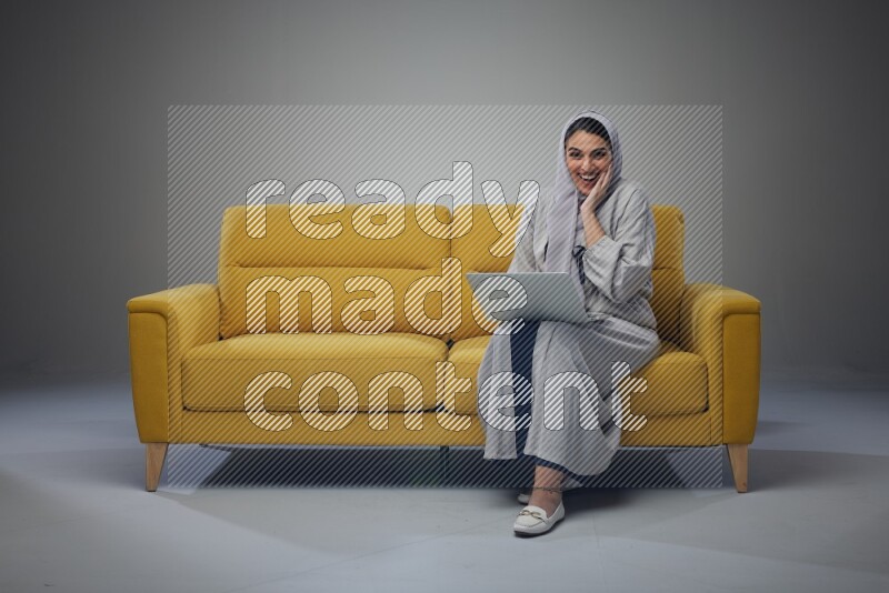 A Saudi woman wearing a light gray Abaya and head scarf sitting on a yellow sofa and using her laptop eye level on a grey background