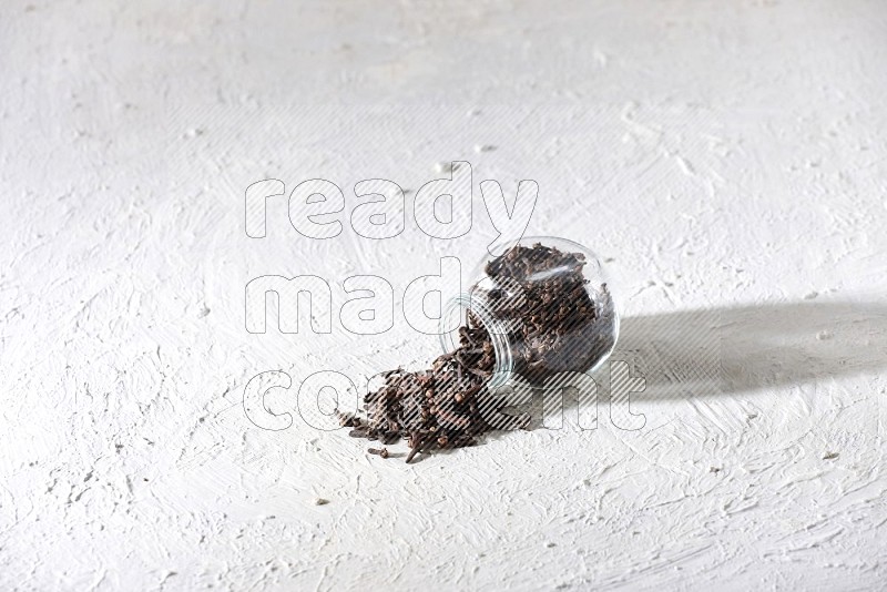 A flipped glass spice jar full of cloves and cloves came out of it on textured white flooring