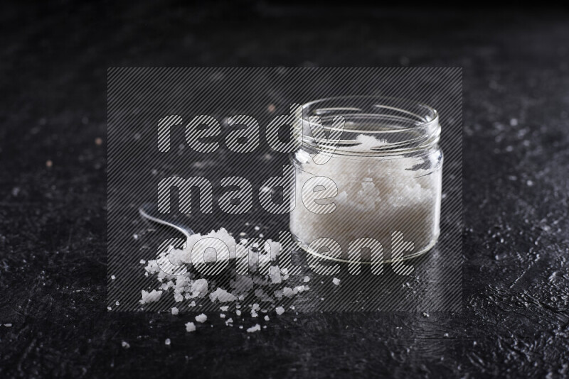 A glass jar full of coarse sea salt crystals on black background