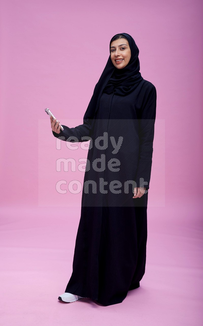 Saudi woman wearing Abaya standing taking selfie on pink background