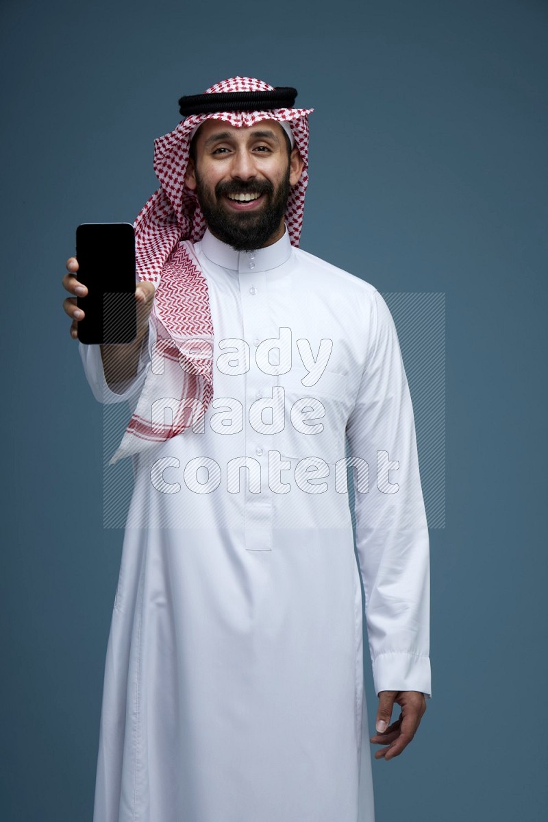 A man showing his phone Screen in a blue background wearing Saudi Thob with Shomag