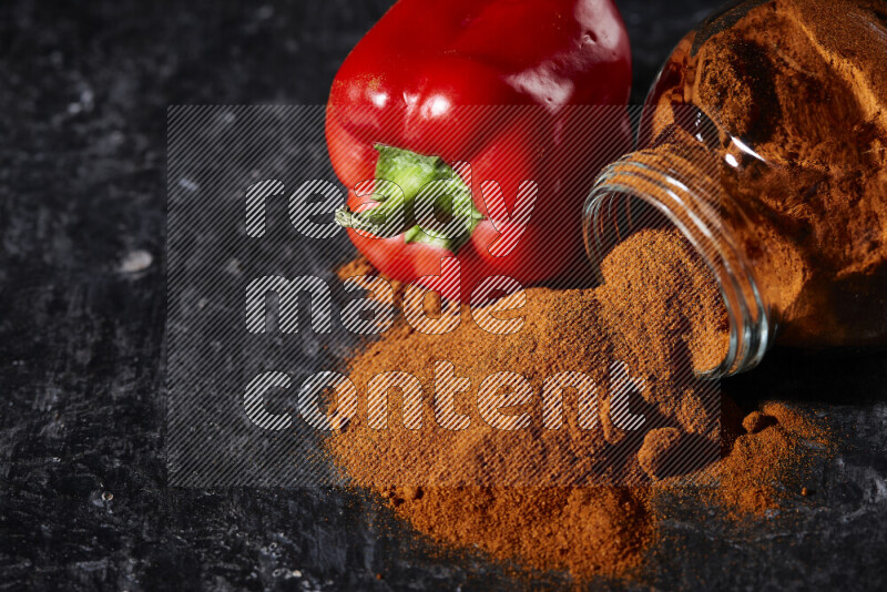 A glass jar full of ground paprika powder flipped with some spilling powder on black background