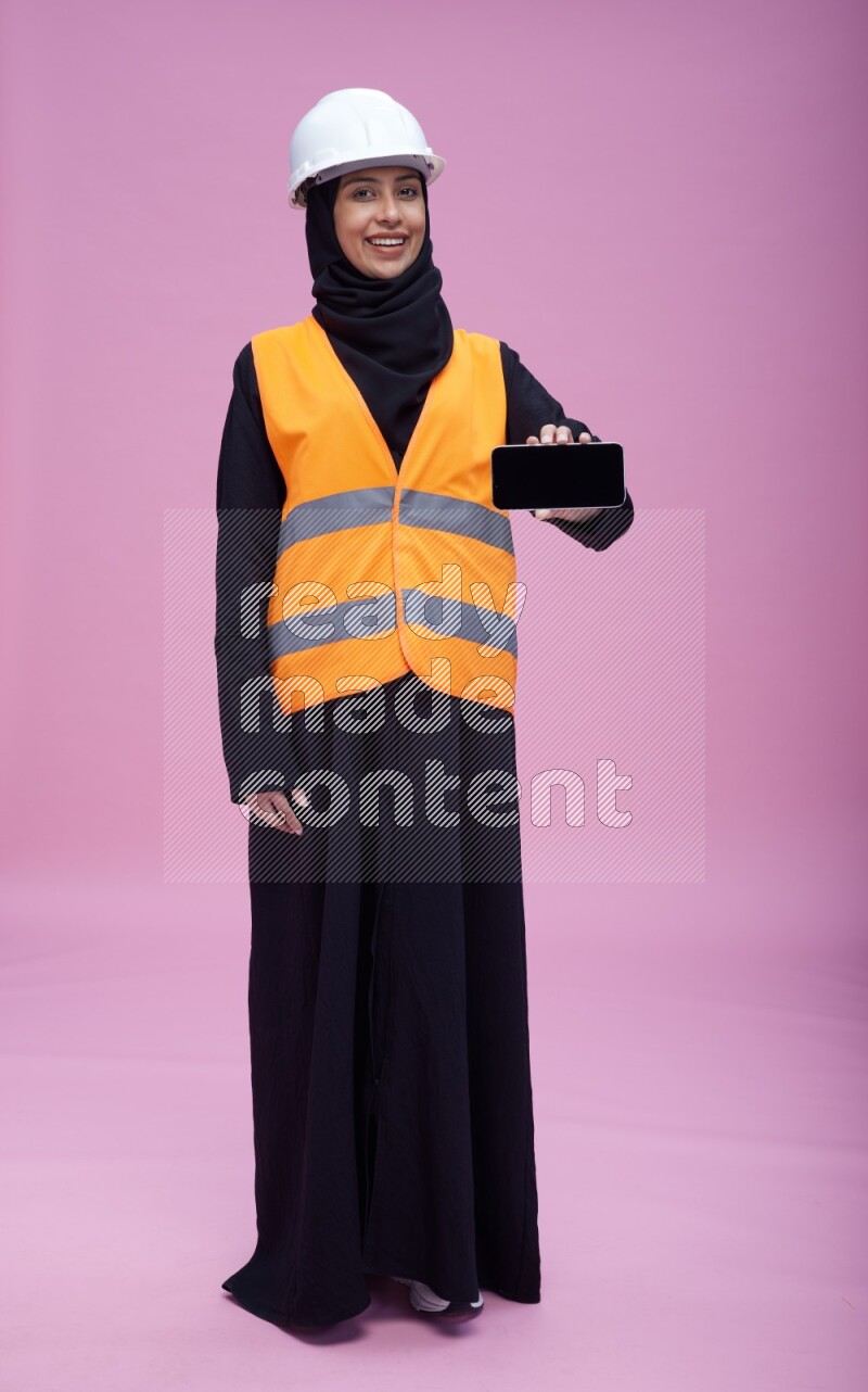 Saudi woman wearing Abaya with engineer vest and helmet standing showing phone to camera on pink background