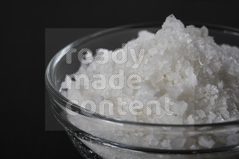 A glass bowl full of coarse sea salt crystals on black background