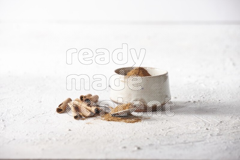 Ceramic beige bowl full of cinnamon powder and a metal spoon with cinnamon sticks next of it on a textured white background
