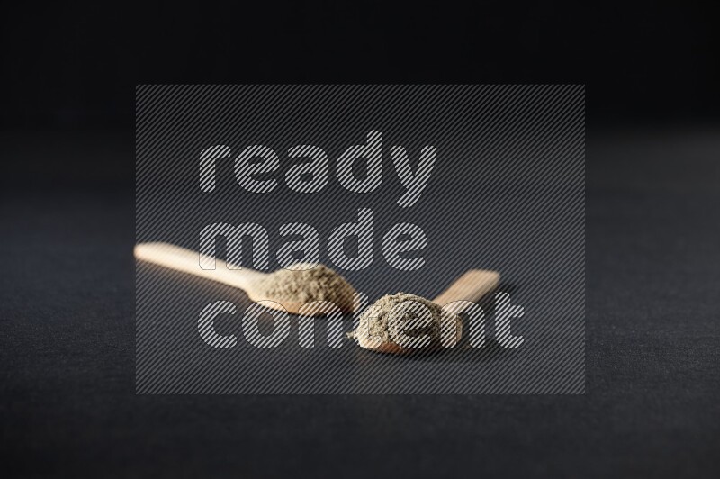 2 Wooden spoons full of cardamom powder on black flooring