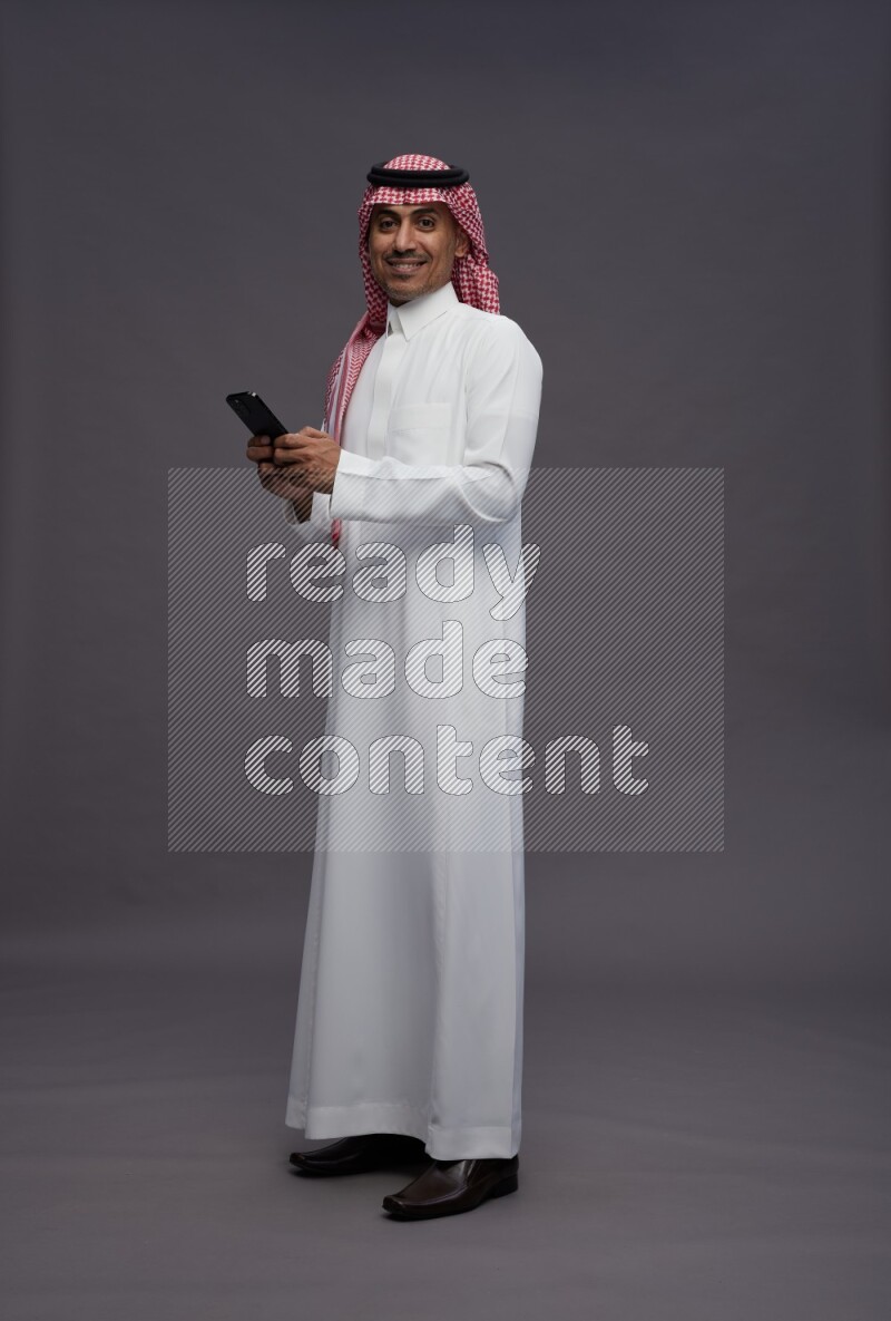 Saudi man wearing thob and shomag standing texting on phone on gray background