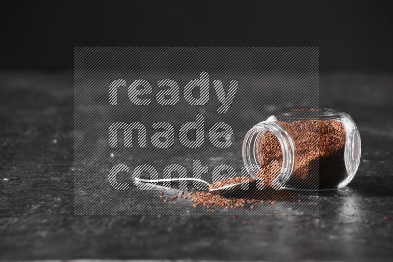 A glass spice jar full of garden cress seeds flipped and seeds spread out with a metal spoon full of the seeds on a textured black flooring