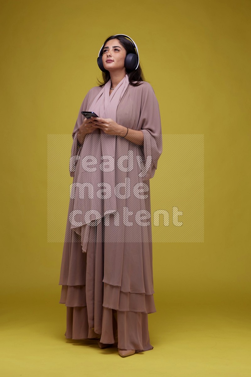 A Saudi woman Listing to Music on a Yellow Background wearing Brown Abaya