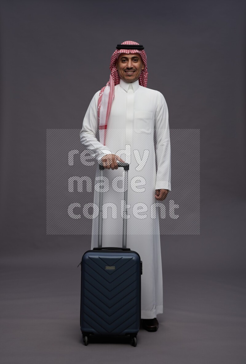 Saudi man wearing thob and shomag standing holding bag on gray background