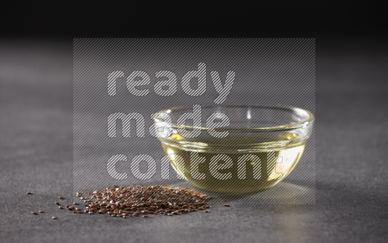 A glass bowl full of flaxseeds oil with some of flaxseeds beside it on a black flooring