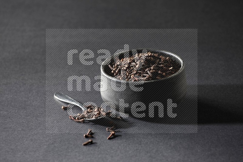 A black pottery bowl full of cloves and a metal spoon next to it on a black flooring