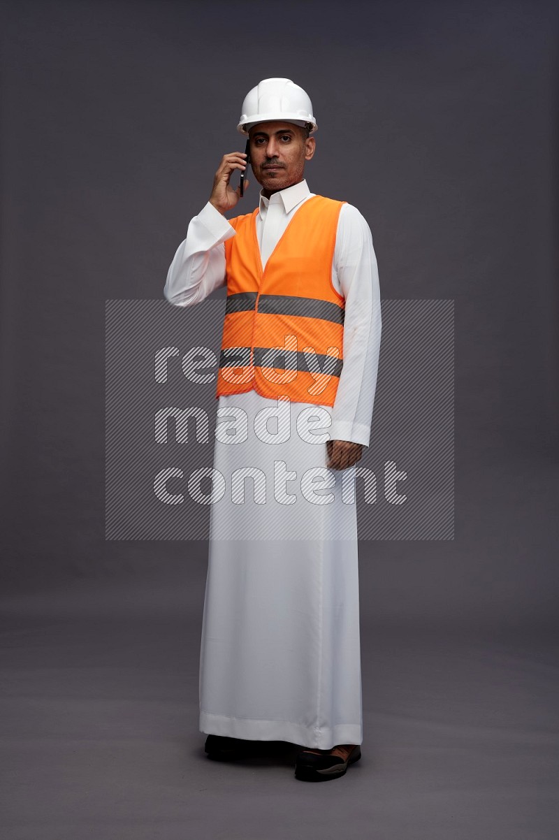 Saudi man wearing thob with engineer vest standing talking on phone on gray background