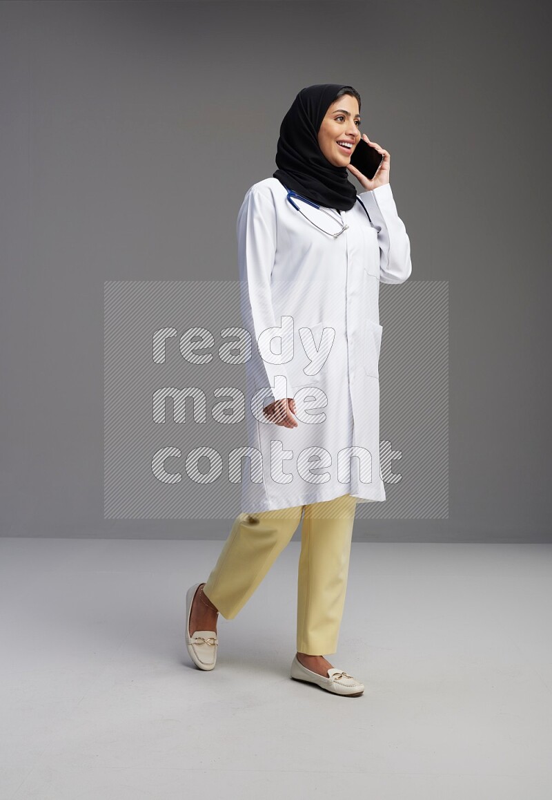 Saudi woman wearing lab coat with stethoscope standing talking on phone on Gray background