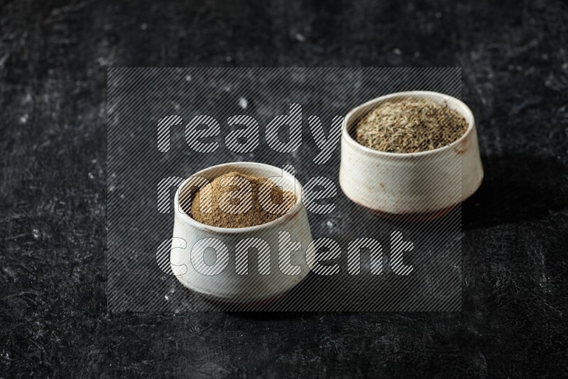 2 beige bowls, one full of cumin powder and the other full of seeds on a textured black flooring