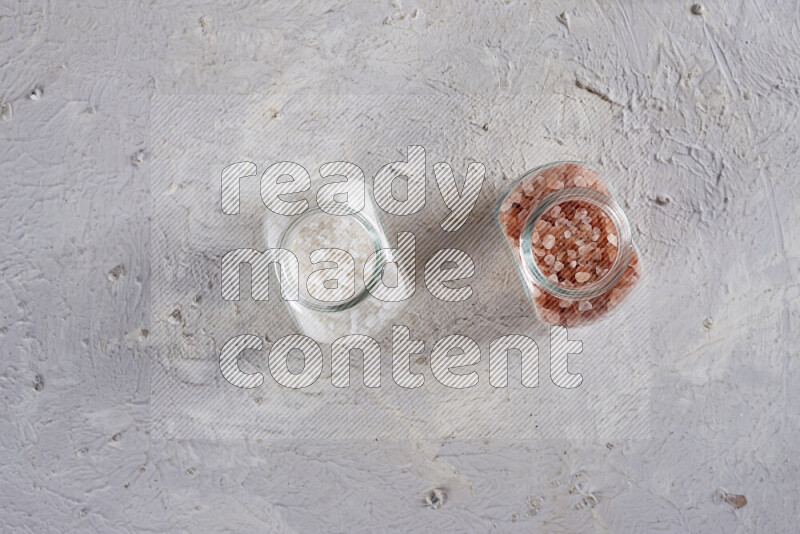 2 glass jars one is filled with coarse himalayan salt and the other with coarse sea salt on white background