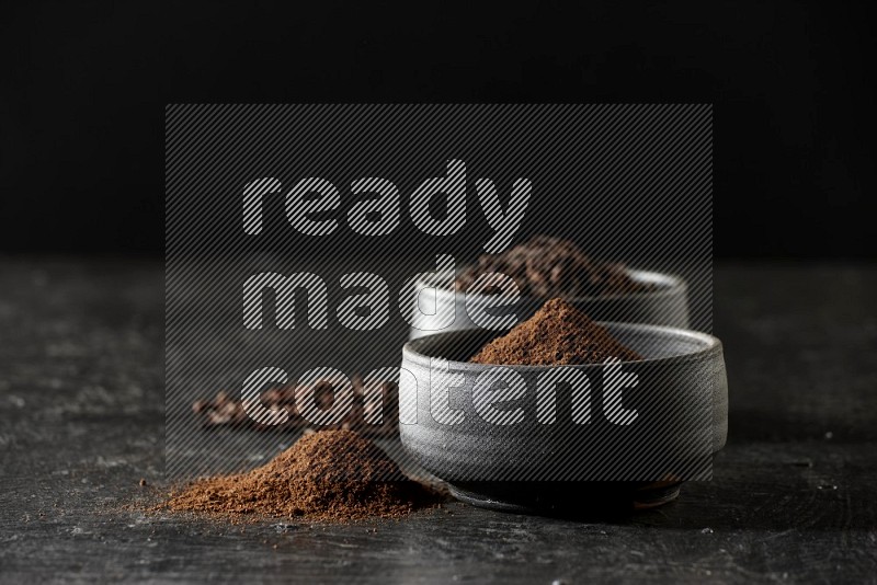 2 Black pottery bowls full of cloves and the other full of cloves powder on textured black flooring