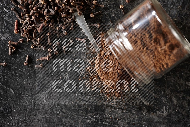 وعاء زجاجي مقلوب وملعقة معدنية ممتلئان ببودرة القرنفل مع حبوب القرنفل المتناثرة على أرضية سوداء