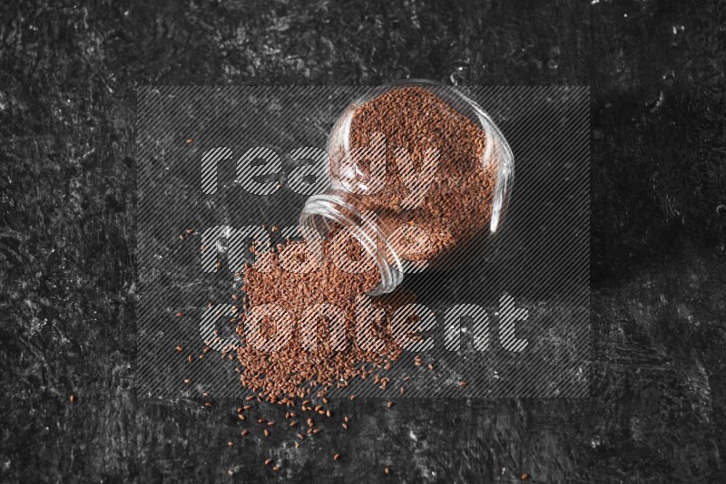 A glass spice jar full of garden cress seeds flipped and seeds spread out a textured black flooring