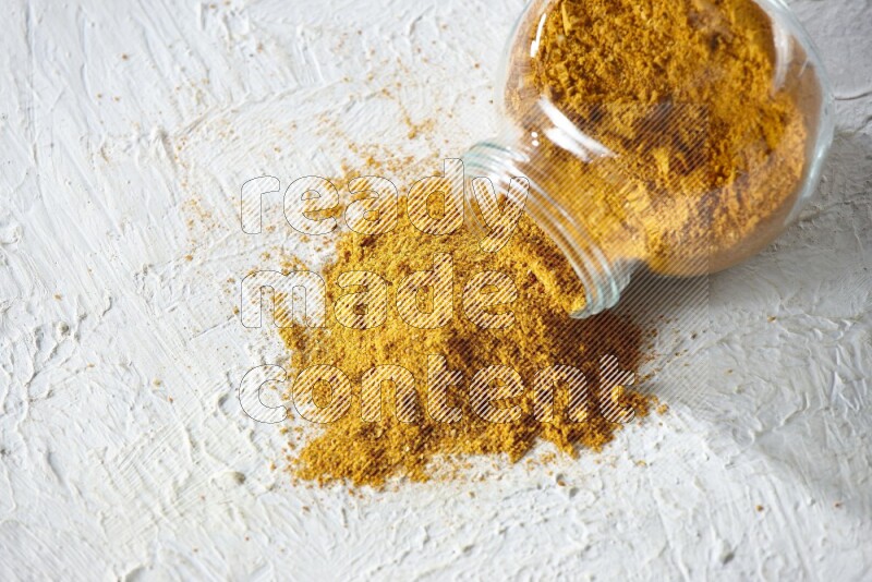 A flipped glass spice jar full of turmeric powder and powder fell out it on textured white flooring