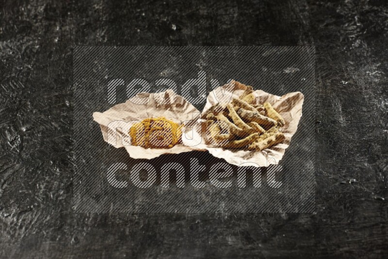 Turmeric powder and dried turmeric whole fingers in 2 crumpled pieces of paper on textured black flooring