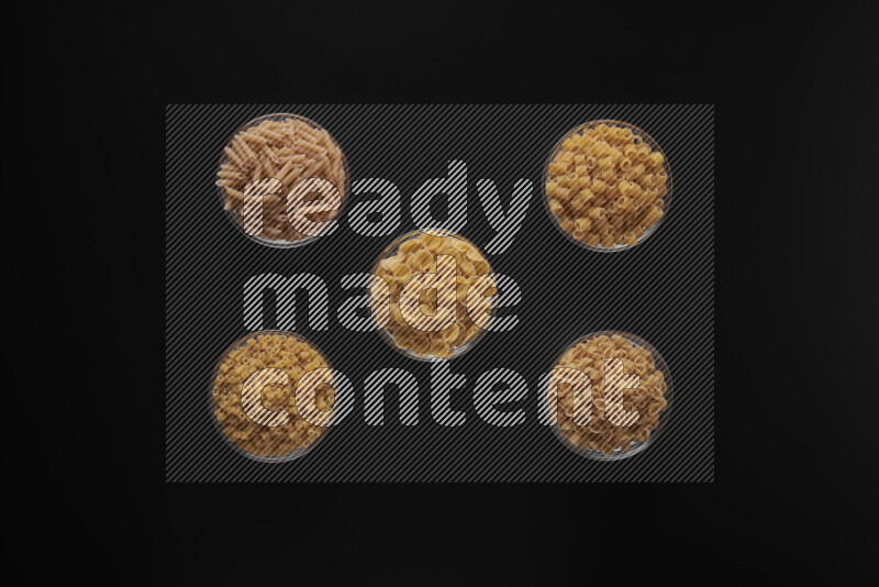 Different pasta types in 5 glass bowls on black background