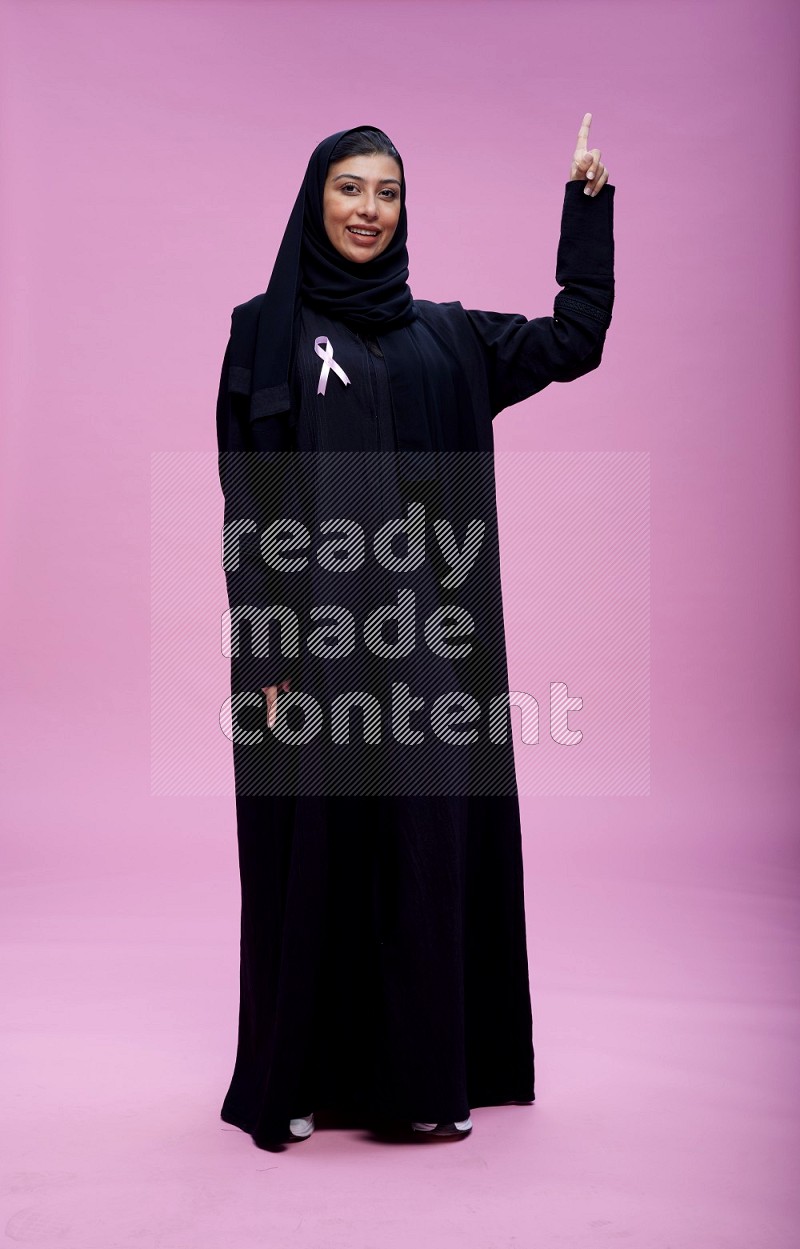 Saudi woman wearing pink ribbon on Abaya standing interacting with the camera on pink background