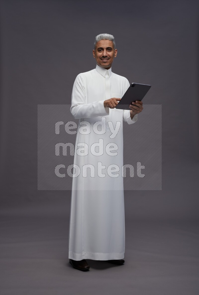 Saudi man wearing thob standing working on tablet on gray background