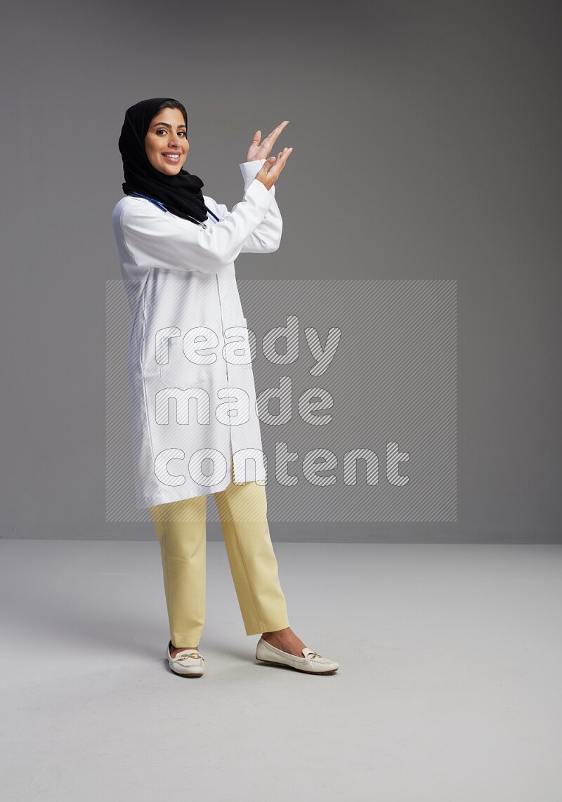 Saudi woman wearing lab coat with stethoscope standing interacting with the camera on Gray background