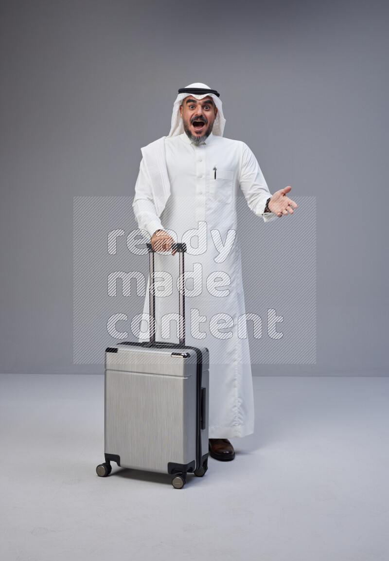 Saudi man wearing Thob and white Shomag standing holding Travel bag on Gray background