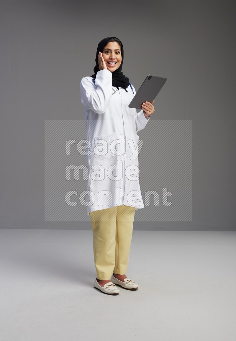 Saudi woman wearing lab coat with stethoscope standing working on tablet on Gray background
