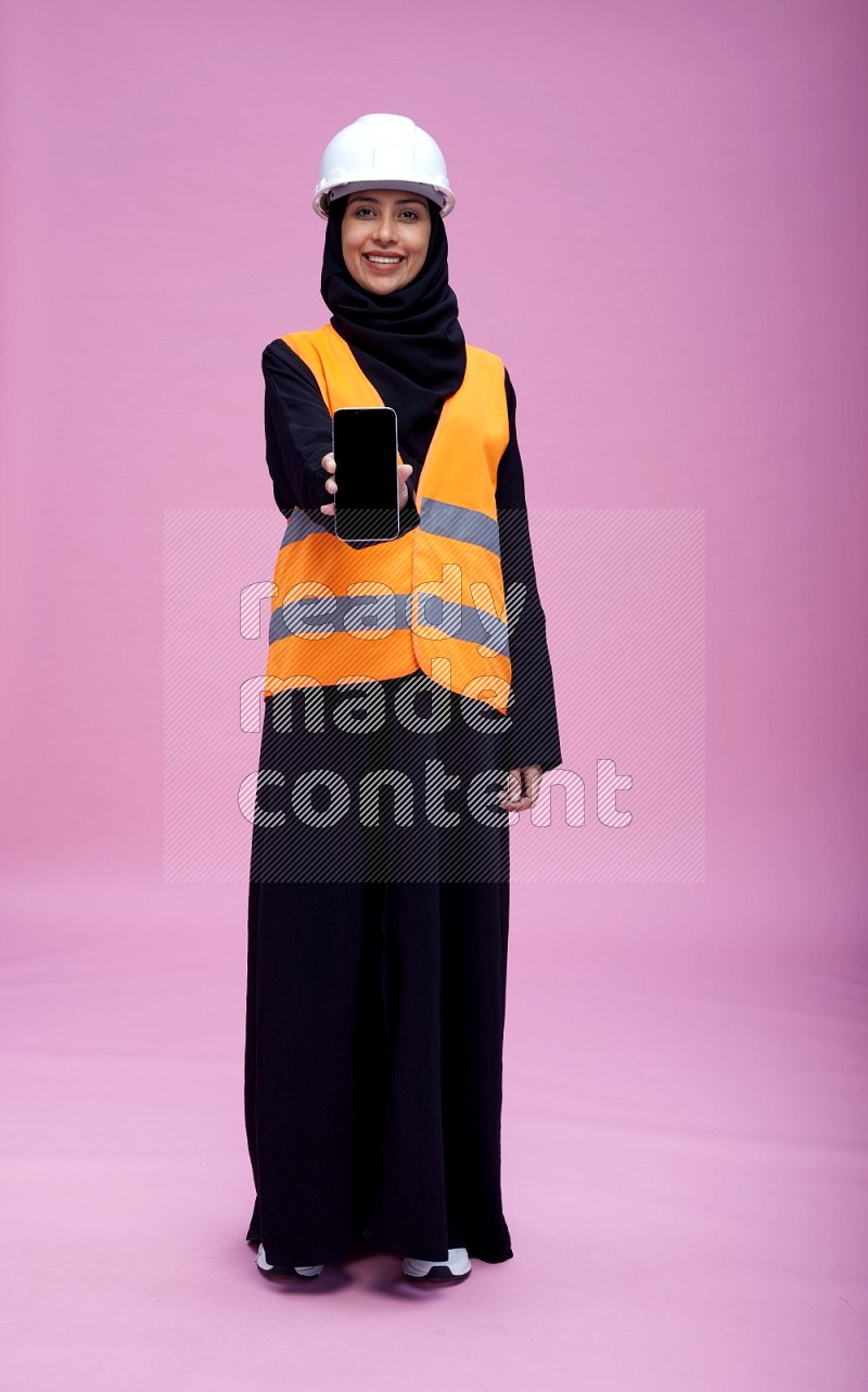 Saudi woman wearing Abaya with engineer vest and helmet standing showing phone to camera on pink background