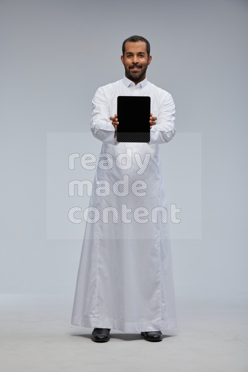 Saudi man wearing Thob standing showing tablet to camera on Gray background