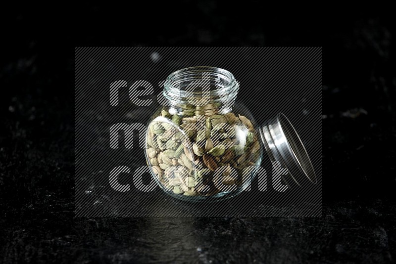A glass spice jar full of cardamom seeds on textured black flooring
