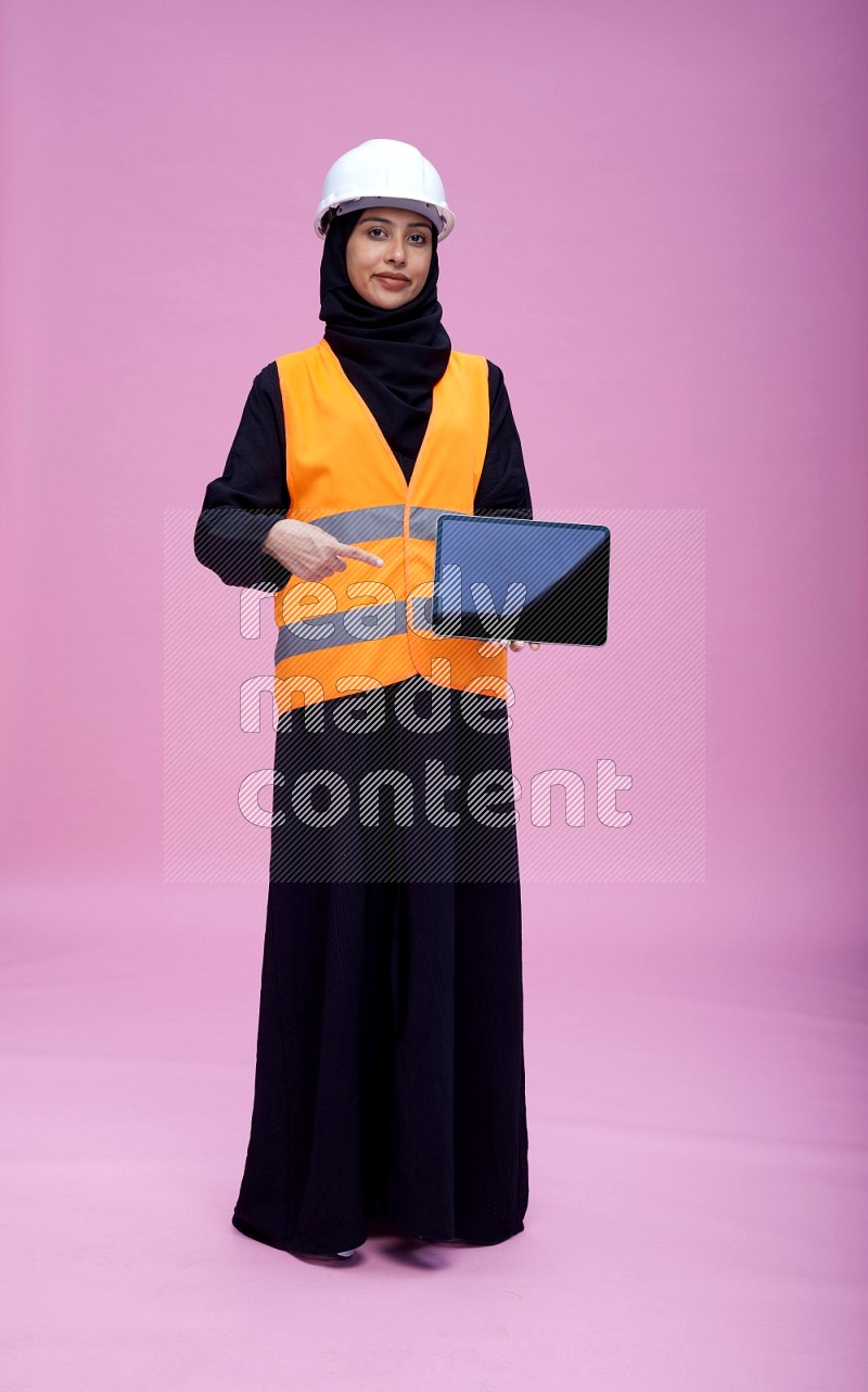 Saudi woman wearing Abaya with engineer vest and helmet standing showing tablet to camera on pink background