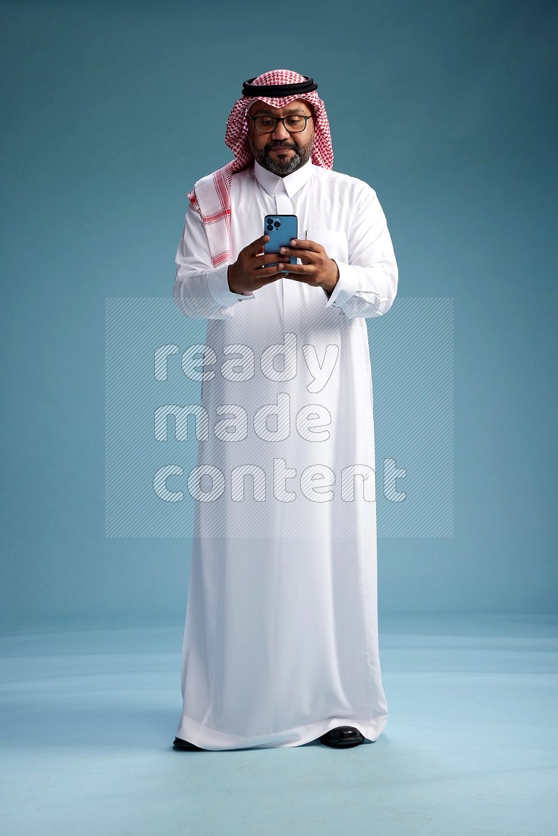 Saudi Man with shimag Standing texting on phone on blue background