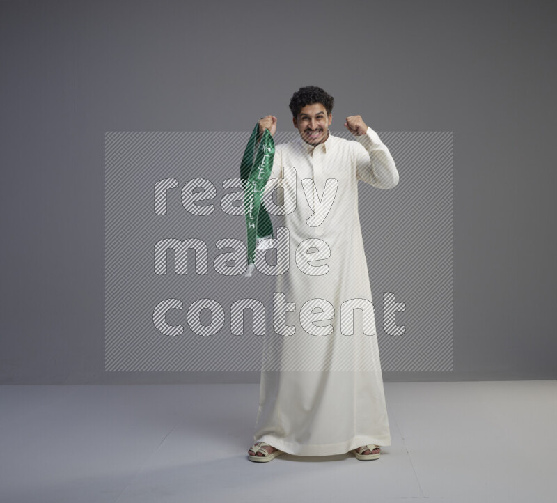 رجل سعودي يرتدي ثوب ابيض ويحمل شال اخضر على خلفية رمادي