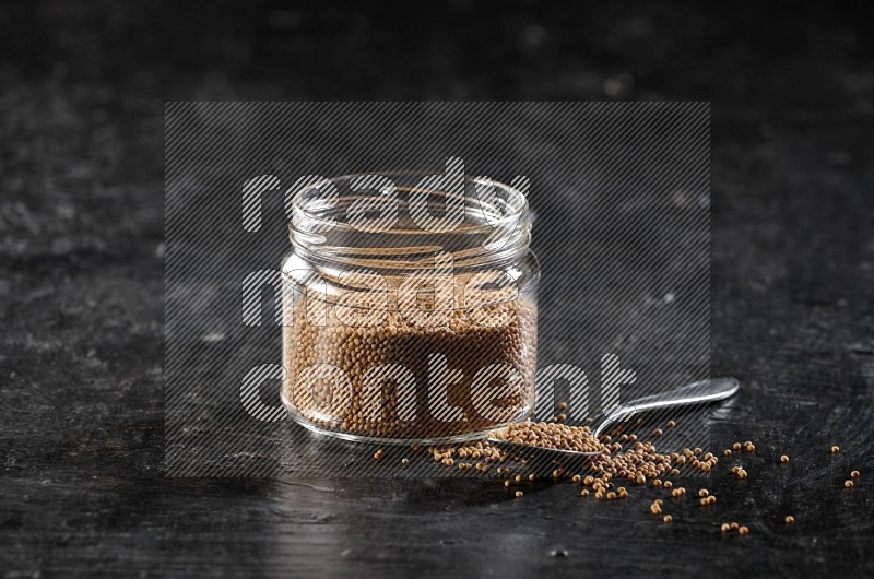 وعاء زجاجي وملعقة معدنية كلاهما ممتلئ ببذور الخردل على أرضية سوداء