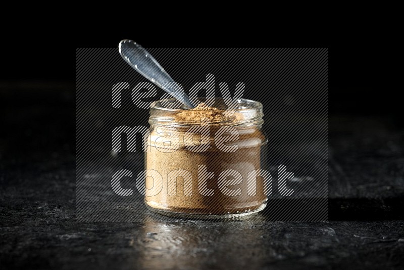 A glass jar and a metal spoon full of allspice powder on a textured black flooring