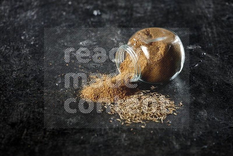 A flipped glass spice jar full of cumin powder with spilled powder and cumin seeds on a textured black flooring