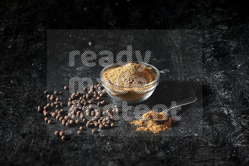 A glass bowl and metal spoon full of allspice powder and whole balls spreaded on a black flooring