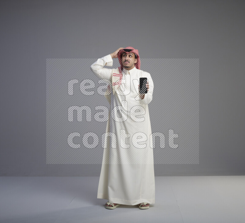 A Saudi man standing wearing thob and red shomag showing phone to camera on gray background