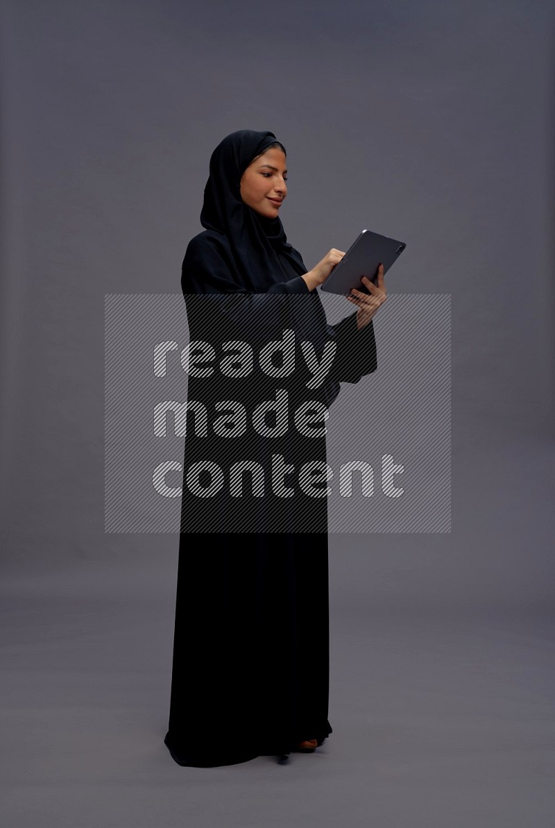 Saudi woman wearing Abaya standing working on tablet on gray background